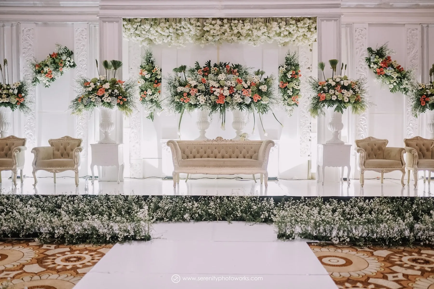 Pelaminan putih dengan rangkaian bunga di ballroom