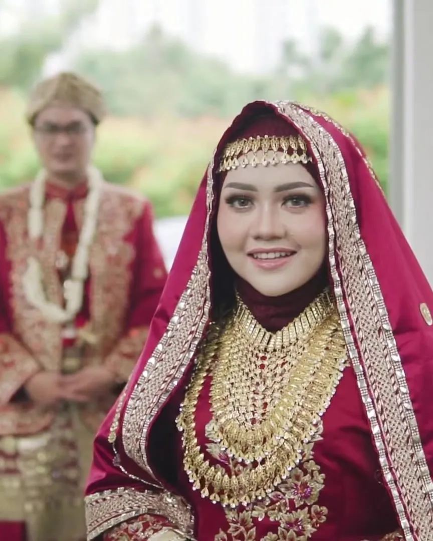 Pengantin wanita berhijab merah marun dengan perhiasan emas, tersenyum ke kamera